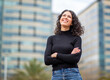 © mimagephotos - woman stands outdoors with arms crossed smiling and looking upward with city buildings in the background.