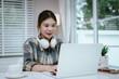 © Worawi - A young woman with long hair wears headphones while working or studying on a laptop at home, enjoying a peaceful, bright, and productive remote learning environment.