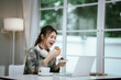 © Worawi - A young woman enjoys coffee and dessert while wearing headphones, working or studying remotely from home in a bright, cozy, and modern digital workspace.