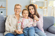 © Studio Romantic - Three generations of smiling women sitting together on couch, looking joyfully at camera. Grandmother, mother and granddaughter resting on sofa, hugging and making family photo in living room at home.