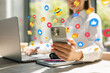 © alones - Woman using smartphone with social media icons floating around, sitting at desk with laptop. Concept of digital communication, online interaction, and modern technology in everyday life.