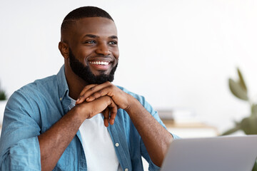 Poster - Portrait of pensive smiling black freelancer sitting at desk in home office, thinking about business strategy while working on laptop computer, looking away with pleased face expression, free space