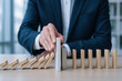 © Chopang.studio - Businessman hand stopping falling wooden dominoes, risk management, crisis prevention and business strategy concept office background professional suit decision making calm and focused mood