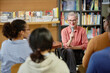 © Seventyfour - Senior Caucasian woman holding microphone speaking to diverse group of young adults and teenagers seated in library setting during session, bookshelves filled with books visible in background