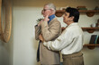 © DragonImages - Senior woman assisting elderly Caucasian husband with choosing right tie, man getting dressed in front of mirror for going out, couple chatting
