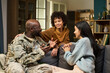 © pressmaster - Black middle aged man in military uniform sitting on couch talking with smiling Asian woman and teenager, family members interacting together in home living room