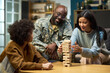 © pressmaster - Caucasian woman and child playing wooden block game with smiling Black middle aged man in military uniform watching at home, family spending quality time together in living room