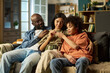 © pressmaster - Black middle aged man in military style clothing sitting on sofa with young adult Caucasian woman and teenage Black boy, family members interacting closely at home
