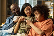 © pressmaster - Caucasian young adult woman, Black middle aged man, and Black child sitting together on sofa, smiling and interacting closely, family spending time at home, military dog tag visible