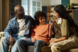 © pressmaster - Black middle aged man sitting with two teenagers on sofa, smiling and interacting together at home, teenage girl showing necklace to teenage boy, family spending time in living room