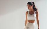 A beautiful woman in white yoga pants is doing morning gym fitness, wearing sportswear and looking at the camera with eyes closed, posing for a photoshoot.