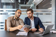 © Liubomir - Two businessmen reviewing a tablet and laptop, discussing business strategies in a modern office setting.