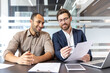 © Liubomir - Two businessmen are reviewing documents in a modern office, discussing plans, and smiling. Collaboration and business are key themes.