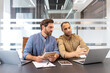© Liubomir - Two diverse businessmen collaborate in a modern office, discussing project details with laptops and documents.