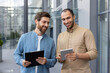 © Liubomir - Two diverse businessmen review a tablet, possibly discussing a project or presenting ideas, smiling in a modern outdoor setting.