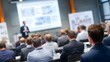 © Pris - A speaker on stage presents to a room full of attendees. The projector shows a slide with images and text. People attentively listen.