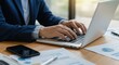 © Khamdani - Business professional typing on laptop amidst financial charts and smartphone.
