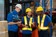 © yuttapon - Three male warehouse workers discussing logistics tasks using digital tablet and clipboard while wearing safety helmets and reflective vests in storage facility surrounded by racks and cardboard boxes