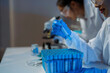 © amnaj - Scientist wearing gloves analyzing blue liquid in test tube with microscope in background in laboratory