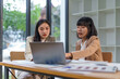 © amnaj - Two Asian businesswomen collaborating and discussing strategies while working on a laptop in a modern office environment