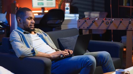 Poster - Adult guy typing on laptop laying on couch in stylish apartment, engaging in freelancing and online work. Black person multitasking efficiently and embracing flexibility of remote job. Camera B.