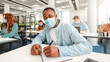 © Prostock-studio - New Rules, Precautions. Portrait of African American male student sitting at table, writing in notebook and looking at camera, wearing disposable medical face mask, afro guy taking notes