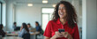 © miss irine - Young smiling mexican community manager wearing red polo shirt in bright white office with colleagues in background. Woman holding phone, using mobile apps. Business communication, social media,