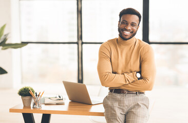 Poster - Confident Businessman. Happy african guy smiling at camera, sitting on desk with crossed arms. Copy space