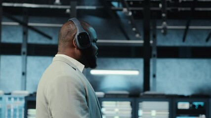Poster - Happy manager listening music, walking in data center with blinking lights on rigs showing system activity. Cheerful man wearing headphones overseeing dimly lit server farm, camera B