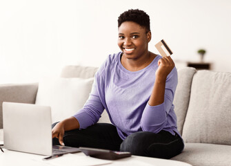 Poster - Making Financial Transaction. Portrait of smiling curvy black lady holding and showing credit card, sitting on the couch with laptop computer at home, looking at camera. Online Money Transfer