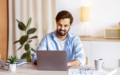Poster - Cheerful Middle Eastern Man Sitting At Desk In Home Office And Working On Laptop. Handsome Arab Male Entrepreneur Using Computer, Enjoying Remote Distance Job Opportunities, Free Space