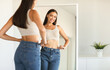 © Prostock-studio - Cheerful young woman posing wearing jeans and smiling to her reflection in mirror after successful weight loss, copy space. Female beauty and style, self confidence, slimming concept