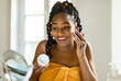 © Prostock-studio - Daily beauty routine. Happy black lady applying moisturising cream on face, looking at magnifying mirror in bedroom, using nourishing skin and smiling, free space