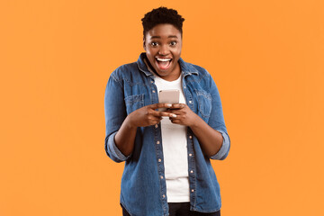 Poster - Great Application. Joyful Black Female Using New Smartphone And Mobile App Posing Smiling To Camera Standing Over Yellow Studio Background. Woman Playing Games On Phone Concept