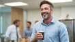 © Attasit - Steaming coffee cup held by male employee in office break room, colleagues laughing nearby.
