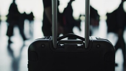 Poster - Stationary black suitcase positioned within bustling airport terminal, passengers moving blurrily across background, capturing modern travel environment and transient moment of waiting