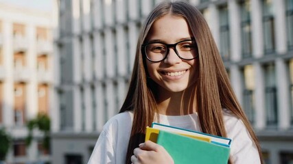 Aufkleber - Portrait of a cheerful teenage student girl with eyeglasses and long hair, wearing a white t shirt and holding colorful books, standing in front of a school, smiling at the camera