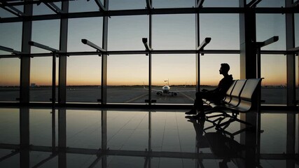 Poster - Young businessman sitting quietly at airport terminal, gazing reflectively through large glass windows at sunset airplane, silhouetted against warm orange sky with soft floor reflection