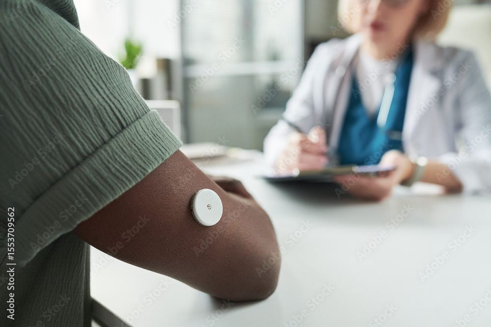 Black young adult man wearing glucose monitor on upper arm consulting with Caucasian middle aged female doctor in medical office, doctor writing notes during diabetes management appointment