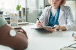 © pressmaster - Middle aged Caucasian female doctor consulting Black adult patient with diabetes, writing notes on clipboard while patient with glucose monitor on arm sitting across medical office desk
