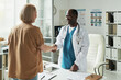 © pressmaster - Middle aged Caucasian woman visiting Black male doctor in medical office, shaking hands across desk, discussing diabetes management with medical charts and documents visible