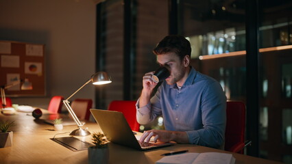 Poster - Overtime manager drinking coffee looking laptop screen at dark office. Tired man