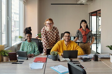 Poster - Group of young adult coworkers collaborating at shared desk with laptops and documents in modern creative coworking space