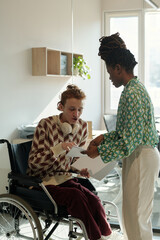 Poster - Young adult Caucasian man sitting in wheelchair discussing documents with young adult Black woman standing beside in creative coworking space