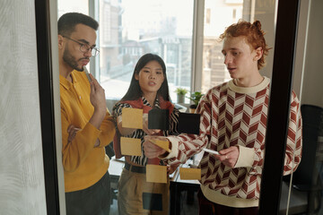 Poster - Caucasian man explaining project details using sticky notes on glass wall while young adult Black man and Asian woman attentively listening