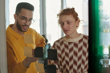 Poster - Young Caucasian man with red hair and Black man with glasses collaborating on creative coworking project, arranging sticky notes on glass wall