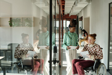Poster - Young adult Black woman standing while talking to young adult Caucasian man sitting in wheelchair with documents in modern coworking office corridor