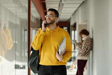 Poster - Young adult man wearing yellow sweater holding documents, talking on phone while walking in modern coworking space, blurred people in background