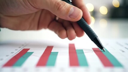 Poster - Financial Chart Analysis with Hand Holding Pen Pointing at Teal Bar on Red Bar Graph With Blurred Lights Background