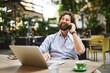 © Stockphotodirectors - A man with curly hair and a beard chats on his phone while typing on his laptop at a cafe. The outdoor setting is vibrant with greenery, creating a relaxed work atmosphere.
